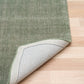 Green area rug corner with stitched edge, rolled back to show the light backing